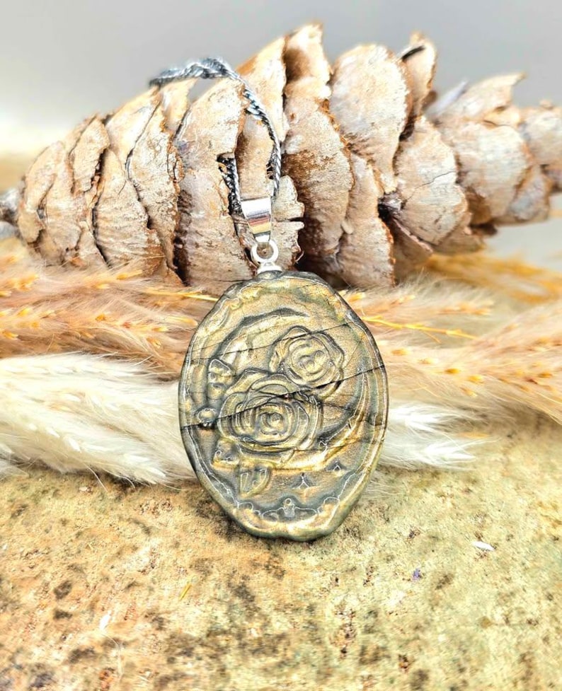 K&ouml;nnte beinhalten: Ein ovaler Anh&auml;nger mit einem geschnitzten Rosendesign in metallischem Gold-Finish. Der Anh&auml;nger h&auml;ngt an einer d&uuml;nnen, dunklen, gedrehten Metallkette. Die Oberfl&auml;che des Anh&auml;ngers weist eine subtile horizontale Textur und Glanz auf.
