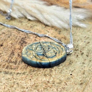 K&ouml;nnte beinhalten: Ein ovaler Anh&auml;nger mit einem geschnitzten Rosenmotiv in Blau- und Goldt&ouml;nen, der an einer silberfarbenen Kette h&auml;ngt. Der Anh&auml;nger hat eine glatte, polierte Oberfl&auml;che mit sichtbarer nat&uuml;rlicher Maserung und metallischem Glanz auf dem Relief.