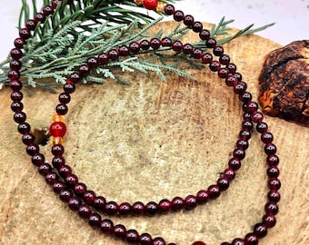 May include: A red garnet mala necklace with gold and red beads. The necklace is laid out on a wooden surface.