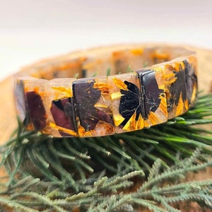 Pode incluir: Um bracelete transparente com inclusões de flores douradas e pretas. O bracelete é feito de várias contas retangulares.