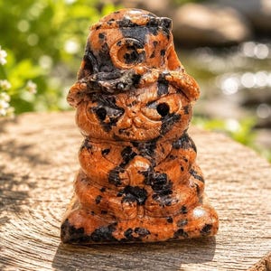 May include: A carved stone figurine depicting a seated creature wearing a wide-brimmed hat. The sculpture is crafted from a mottled orange and black mineral, featuring deep-set eyes, a small nose, and hands clasped together in a meditative pose.