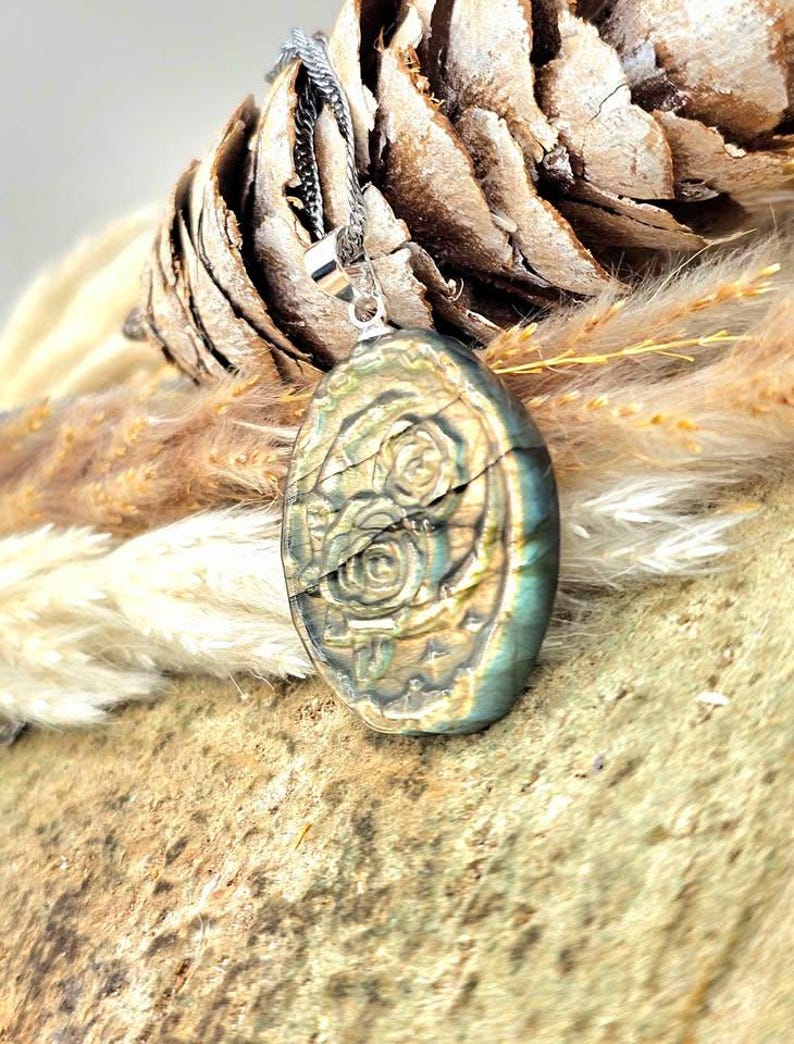 K&ouml;nnte beinhalten: Ein ovaler Labradorit-Anh&auml;nger mit einer eingravierten Blumenzeichnung von zwei Rosen. Der Stein zeigt irisierende Blitze in Blau-, Gold- und Gr&uuml;nt&ouml;nen. Er h&auml;ngt an einer silberfarbenen Metall&ouml;se und weist eine polierte Oberfl&auml;che auf.