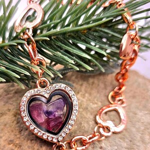 May include: Rose gold bracelet featuring a heart-shaped locket filled with purple stones, surrounded by clear crystals. The bracelet has a chain design with heart-shaped and oval links. The background includes green pine needles.
