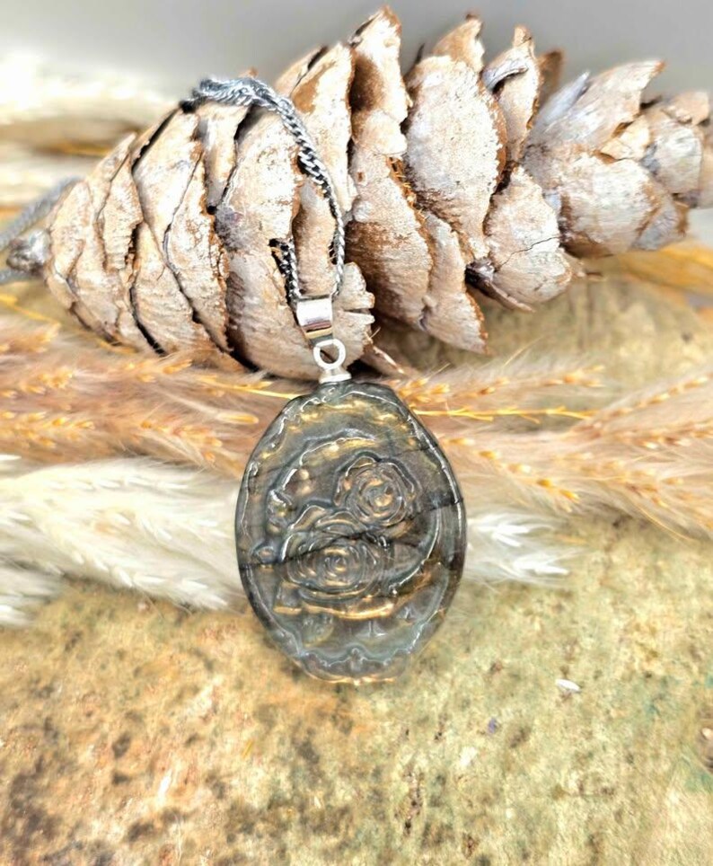 K&ouml;nnte beinhalten: Ein ovaler Anh&auml;nger mit einem geschnitzten Rosenmotiv auf einem dunklen, schillernden Stein. Der Anh&auml;nger h&auml;ngt an einer silberfarbenen Kette und zeigt einen metallischen Glanz sowie filigrane florale Details auf der Oberfl&auml;che des Steins.