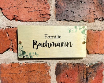 May include: A gold-coloured metal sign with a green leafy design and the text "Familie Bachmann" on a brick wall.