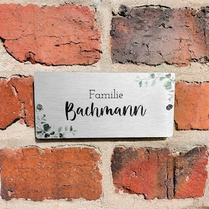 Pode incluir: Uma placa de metal prateada com um design de folhas verdes e o texto "Familie Bachmann" em uma parede de tijolos.
