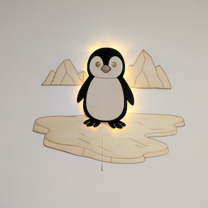 Può includere: Applique a forma di pinguino in legno. Il pinguino è bianco e nero, in piedi su un iceberg di legno colorato. Due ritagli di montagna in legno sono dietro il pinguino. Un cordoncino pende sotto.