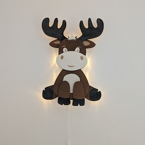 May include: A wooden moose-shaped wall light, featuring a brown body, black antlers, and a white face. The light emits a warm glow, and a white cord hangs below. The moose is designed to sit on a wall.