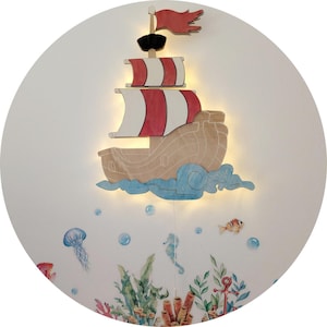 May include: A wooden pirate ship wall decoration with red and white sails, a black mast, and a red flag. The ship is mounted on a white wall with underwater-themed decals, including a jellyfish, seahorse, and fish.