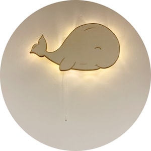 May include: A wooden whale-shaped wall light. The light emits a soft, warm glow, and the whale is a light beige colour. The fixture is mounted on a white, circular background. A thin cord hangs from the bottom of the light.