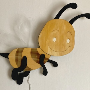 Può includere: Un'ape di legno con un viso sorridente e ali nere. L'ape è dipinta con strisce marroni e gialle.