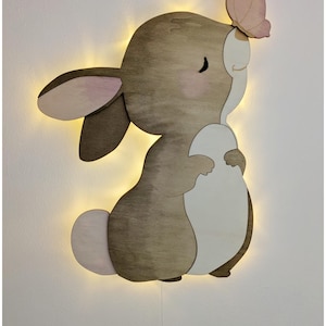 Peut inclure: Lampe murale en forme de lapin en bois avec un papillon. La lumière a un corps marron, des accents roses et un ventre blanc. Le lapin tient une forme blanche et a une lumière sur les bords. Un papillon rose est posé sur sa tête.