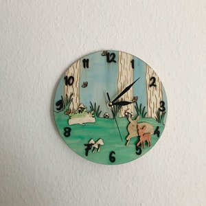 Può includere: Un orologio in legno con una scena di foresta che presenta un cielo blu, erba verde, alberi marroni e due cervi. Il quadrante dell'orologio ha numeri e lancette neri.
