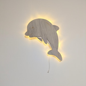 May include: A wooden dolphin-shaped wall light fixture. The light is a neutral gray color with a warm yellow glow emanating from behind the dolphin. A white cord hangs from the bottom.