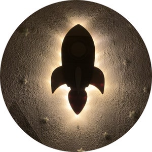 Può includere: Un'illuminazione notturna a forma di razzo in legno bianco e marrone con una luce bianca brillante intorno. Il razzo è su uno sfondo bianco con piccole stelle bianche.