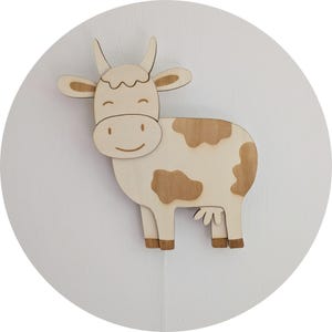 Puede incluir: Decoración de pared con forma de vaca de madera. La vaca tiene un cuerpo de color claro con manchas y detalles marrones. Presenta una cara sonriente, cuernos y orejas. La vaca está montada sobre un fondo circular blanco.