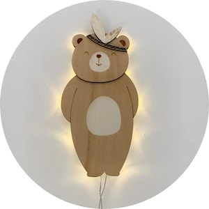 Puede incluir: Una luz nocturna de madera en forma de oso con un tocado de plumas. El oso es marrón con la barriga blanca y la nariz marrón claro. La luz está encendida e ilumina al oso.