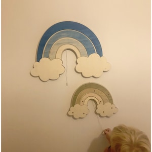 Peut inclure: Deux arcs-en-ciel en bois avec des nuages accrochés à un mur blanc. Le plus grand arc-en-ciel est bleu et blanc, et le plus petit est vert, marron et blanc.