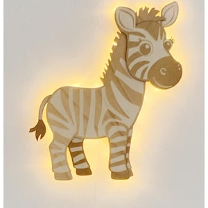 Puede incluir: Una lámpara de pared de madera con forma de cebra, con un brillo amarillo cálido. La cebra tiene rayas marrones claras y crema, con una melena, cola y orejas detalladas. Ideal para una habitación infantil.