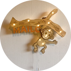 Op de afbeelding: Een houten nachtlampje in de vorm van een aap die aan een tak hangt met een naam, geboortedatum en afmetingen. De tak heeft kleine lampjes die erin zijn ingebed. De aap is bruin met een lichtbruin gezicht en oranje details. De tekst op de tak luidt "21.02.2012 - 3690g - 50cm" en de naam "MAILO" staat onder de tak.