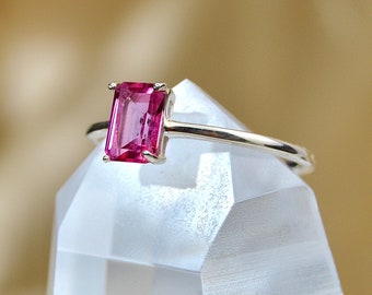 May include: A pink gemstone ring with a rectangular cut stone set in a thin silver band. The ring is displayed on a clear crystal stand.