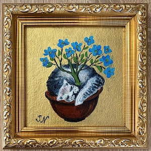 May include: A framed painting featuring a sleeping cat nestled in a terracotta pot, with a tree of blue flowers growing from it. The artwork is set against a gold background and framed in an ornate gold frame.