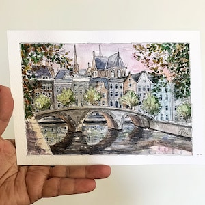 May include: Watercolor painting of a cityscape with a bridge over a canal. The buildings are in shades of gray and brown, with some green trees. The sky is a light pink and blue.