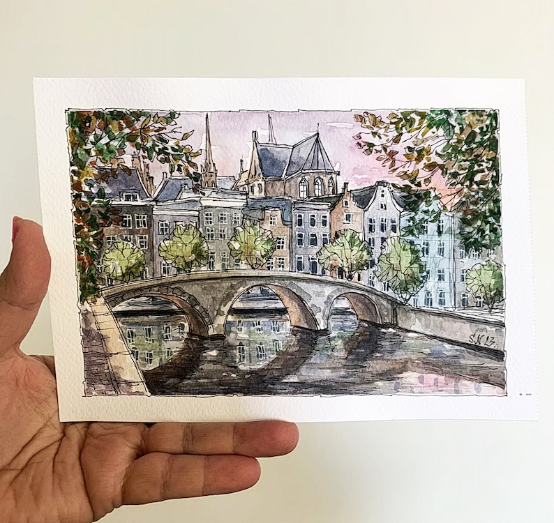 May include: Watercolor painting of a cityscape with a bridge over a canal. The buildings are in shades of gray and brown, with some green trees. The sky is a light pink and blue.