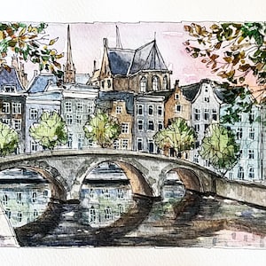 May include: Watercolor painting of a bridge over a canal in a European city. The bridge is made of stone and has three arches. There are buildings on both sides of the canal, and trees lining the banks. The sky is a soft pink and blue.