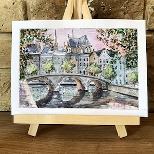 May include: Watercolor painting of a bridge over a canal in a European city. The buildings are brick and have a traditional style. The sky is a soft pink and the water is a dark blue. The painting is signed "SK12."