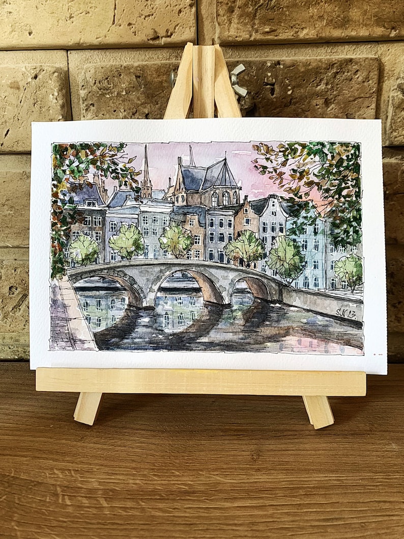 May include: Watercolor painting of a bridge over a canal in a European city. The buildings are brick and have a traditional style. The sky is a soft pink and the water is a dark blue. The painting is signed "SK12."