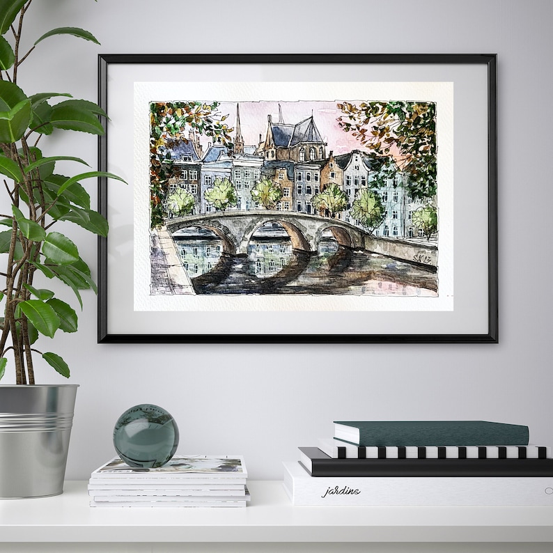 May include: Watercolor painting of a bridge over a canal in a European city. The painting features buildings with red tile roofs, trees, and a reflection of the bridge in the water.