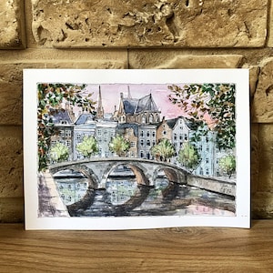 May include: Watercolor painting of a bridge over a canal in a European city. The bridge is made of stone and has three arches. The canal is lined with buildings, some of which have red tile roofs. The sky is a soft pink and blue.