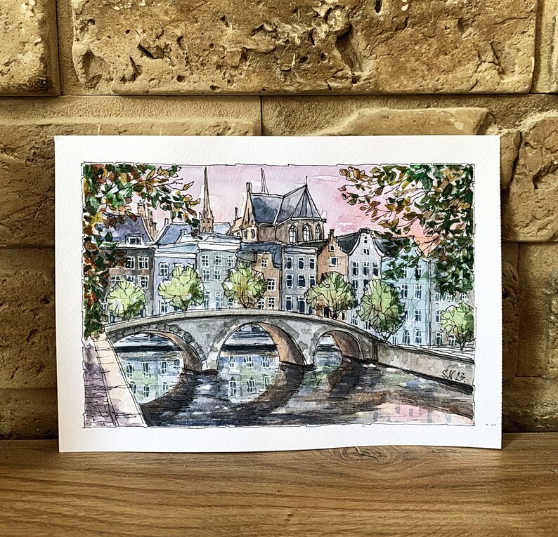 May include: Watercolor painting of a bridge over a canal in a European city. The bridge is made of stone and has three arches. The canal is lined with buildings, some of which have red tile roofs. The sky is a soft pink and blue.