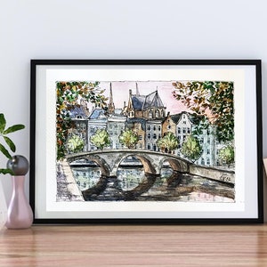 May include: Watercolor painting of a bridge over a canal in a European city. The buildings are in the background, and the sky is a soft pink. The painting is framed in black.