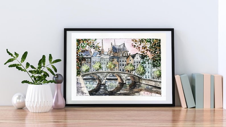 May include: Watercolor painting of a bridge over a canal in a European city. The buildings are in the background, and the sky is a soft pink. The painting is framed in black.