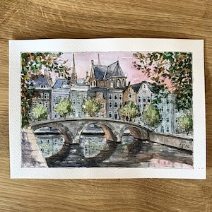May include: Watercolor painting of a bridge over a canal in a European city. The buildings are a mix of brick and stone, with a few trees in the foreground. The sky is a soft pink and blue, with a few clouds.