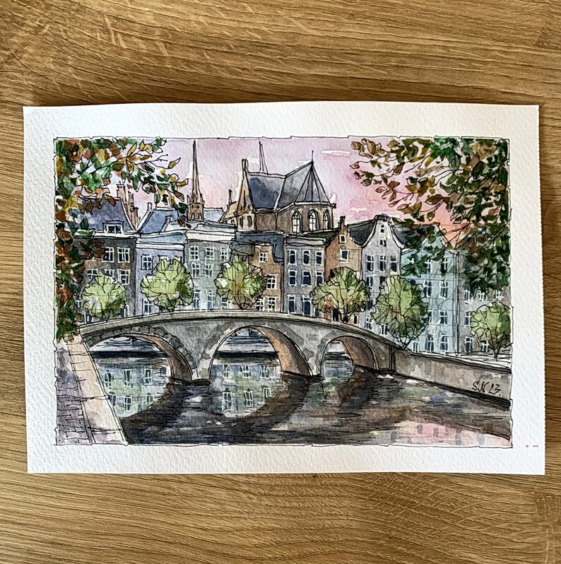May include: Watercolor painting of a bridge over a canal in a European city. The buildings are a mix of brick and stone, with a few trees in the foreground. The sky is a soft pink and blue, with a few clouds.