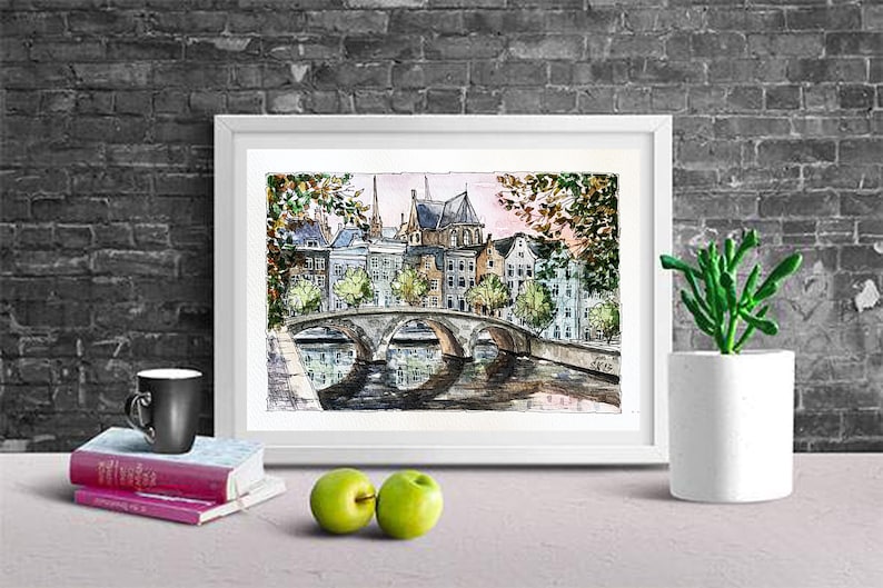 May include: Watercolor painting of a bridge over a canal in a European city. The buildings are in the background and have a light brown color. The bridge is made of stone and has a light gray color. The water is a light blue color. The painting is framed in a white frame.
