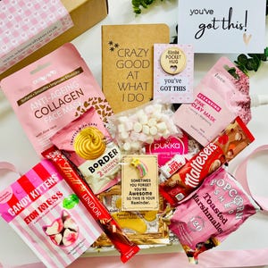 May include: A gift box containing an assortment of items including a pink anti-ageing collagen face mask, a notebook with the text CRAZY GOOD AT WHAT I DO, a pink clay mask, a bag of marshmallows, a Lindt chocolate bar, and Candy Kittens sweets.
