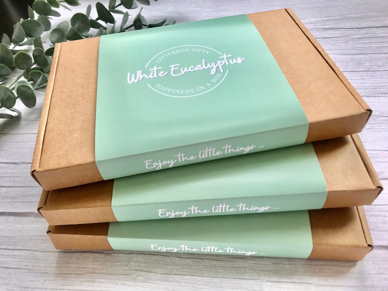 May include: Three brown cardboard boxes stacked on top of each other. The top box has a light green label with the text "LETTERBOX GIFTS White Eucalyptus HAPPINESS IN A BOX" and the text "Enjoy the little things..." printed in white. The middle and bottom boxes have the same light green label with the text "Enjoy the little things..." printed in white.