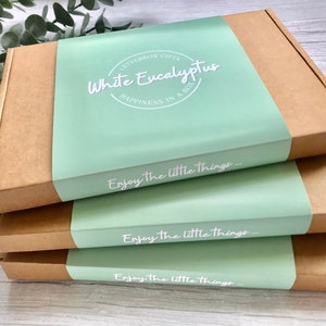 May include: Three brown cardboard boxes stacked on top of each other. The top box has a light green label with the text "LETTERBOX GIFTS White Eucalyptus HAPPINESS IN A BOX" and the text "Enjoy the little things..." printed in white. The middle and bottom boxes have the same light green label with the text "Enjoy the little things..." printed in white.