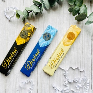 May include: Three Divine chocolate bars on a white wooden surface. The bars are wrapped in black, blue, and white paper with gold lettering. The text on the bars reads "Divine", "70% Smooth Dark Chocolate", "Smooth Milk Chocolate", and "Creamy White Chocolate".