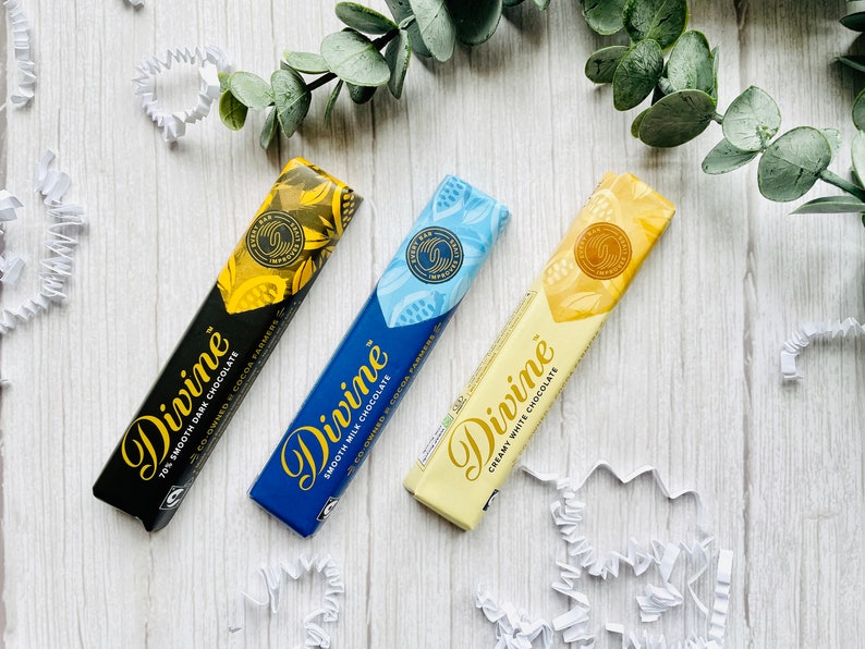 May include: Three Divine chocolate bars on a white wooden surface. The bars are wrapped in black, blue, and white paper with gold lettering. The text on the bars reads "Divine", "70% Smooth Dark Chocolate", "Smooth Milk Chocolate", and "Creamy White Chocolate".