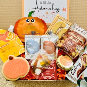 May include: A gift box labelled a little Autumn hug containing a pumpkin-shaped face mask, orange peel-off mask, eye masks, Popcorn Shed cinnamon roll popcorn, a Galaxy chocolate bar, a pumpkin cookie, a small candle, and a bag of gummy sweets.