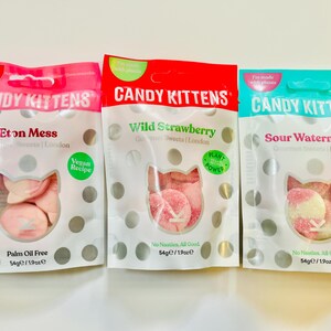 May include: Three individually packaged bags of vegan candy kittens. The bags are pink, red, and blue. The flavors are Eton Mess, Wild Strawberry, and Sour Watermelon. Each bag has a white polka dot pattern and a picture of pink candy kittens.