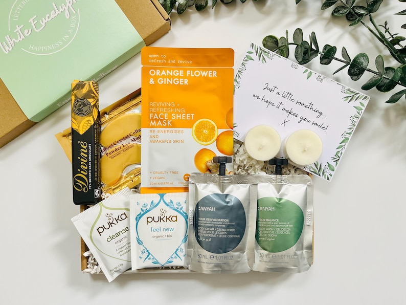 May include: A gift box filled with self-care items, including face masks, bath products, tea, and candles. The box is decorated with shredded paper and a handwritten note that says "Just a little something... we hope it makes you smile!"