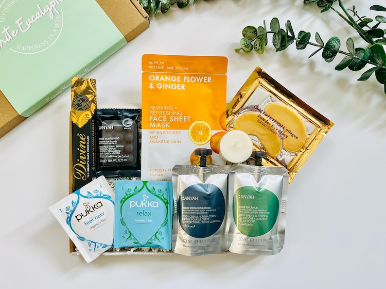 May include: A gift box filled with self-care items, including face masks, eye masks, tea bags, and bath products. The box is decorated with green and white paper and includes the text "Happiness in a Box".