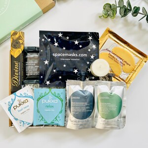 May include: A gift box filled with self-care items, including a chocolate bar, tea bags, face masks, and a candle. The box is decorated with a green and white label that says "Happiness in a Box".