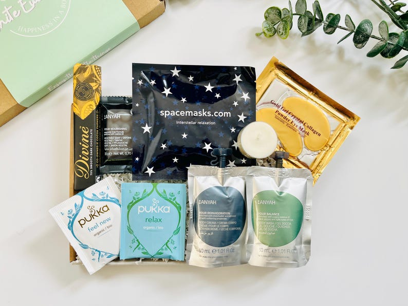 May include: A gift box filled with self-care items, including a chocolate bar, tea bags, face masks, and a candle. The box is decorated with a green and white label that says "Happiness in a Box".